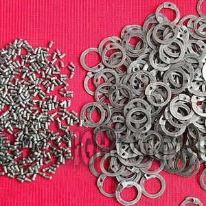 May include: A collection of small, silver-colored metal fasteners and retaining rings are scattered on a bright red surface. The fasteners are cylindrical, while the rings have a circular shape with a flat edge.
