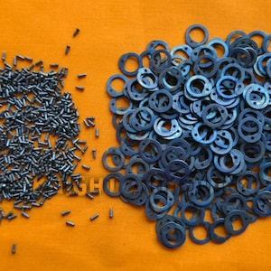May include: An overhead shot displays a collection of small, dark metal screws and a pile of blue-toned metal washers with circular and semi-circular cutouts. The items are arranged on an orange surface.