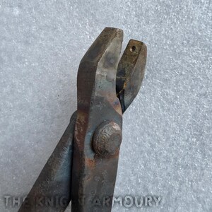 May include: A pair of heavy-duty metal pincers with a dark, weathered finish and a central pivot bolt. The tool features a textured, oxidized surface and a small circular opening on one jaw. The text THE KNIGHT ARMOURY is visible at the bottom.