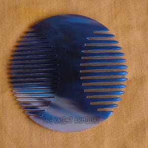 Könnte beinhalten: Ein blauer, kreisförmiger Kamm mit gleichmäßig verteilten Zähnen. Der Kamm hat eine reflektierende Oberfläche und der Text "THE KNIGHT ARMOURY" ist unten aufgedruckt. Der Kamm liegt auf einem beigefarbenen Stoffhintergrund.