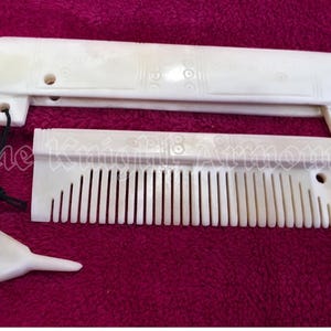 Puede incluir: Juego de peine y estuche de color marfil. El peine tiene dientes finos y es rectangular. El estuche es una pieza larga y curvada con detalles decorativos. Una pequeña herramienta de color marfil está unida con un cordón negro. El texto "The Knight Armory" es visible.