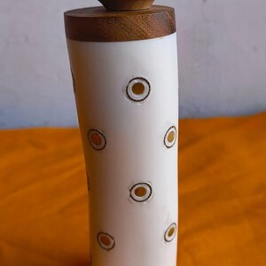 May include: A decorative bottle stopper with a white ceramic body and a carved wooden top. The body has a pattern of brown and gold circles. The top is a carved wooden sphere. The text "THE KNIGHT ARMOURY" is at the bottom.