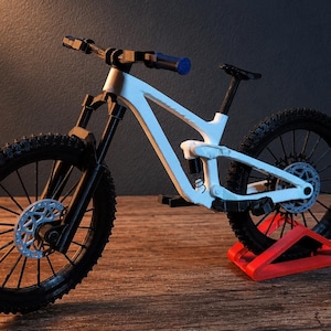 May include: A white and black mountain bike model with a blue handlebar grip. The bike has black tires, a black seat, and a red stand. The bike's frame is white, and the wheels have silver disc brakes.