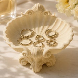 Könnte beinhalten: Eine cremefarbene, muschelförmige Schmuckschale mit mehreren goldenen Ringen. Die Schale ist aufwendig verziert und steht auf einem Sockel. Eine Parfümflasche und ein Strauß weißer Blumen befinden sich im Hintergrund.