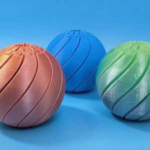 May include: Three spherical objects in shades of copper, blue, and green are arranged against a blue backdrop. Each sphere has a spiral design carved into its surface, creating a textured appearance. The objects appear to be 3D-printed.