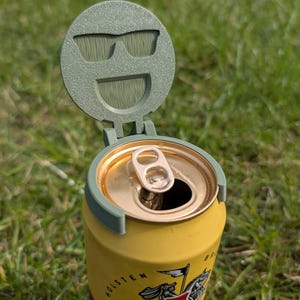 Op de afbeelding: Een gele Holsten bierblikje met een groen deksel met een zonnebril en smiley ontwerp. Het blikje is open, waardoor het lipje en de gouden binnenkant zichtbaar zijn. Het blikje staat op een bed van groen gras.