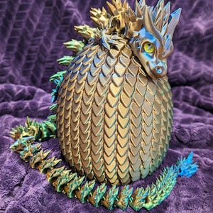 May include: A 3D-printed dragon figurine with a detailed, scale-like body in gold and teal hues. The dragon has a spiky crest and a long, articulated tail. The figurine is resting on a purple, textured surface.