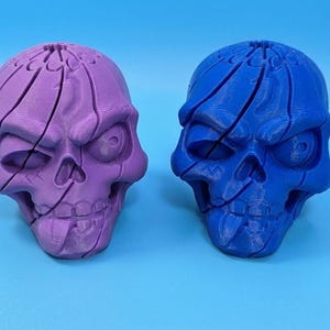 May include: Two 3D-printed skull sculptures, one purple and one blue, sit against a blue background. The skulls have a segmented design, with visible lines across the surface. The eyes are large and menacing, giving them a spooky appearance.