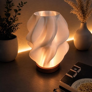 May include: A white, sculptural table lamp with a swirling design, resting on a wooden base. The lamp emits a warm glow, illuminating the surrounding objects, including a potted plant, a vase, and a book.