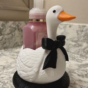 May include: A white goose-shaped soap dispenser holder featuring a black bow around its neck and a black circular base. The holder contains a clear plastic pump bottle filled with pink liquid soap, with a bright orange beak on the goose figure.