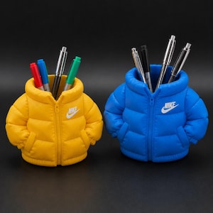 May include: Two pen holders shaped like miniature puffer jackets, one yellow and one blue, each with the Nike logo. The yellow holder contains red, blue, and green pens, while the blue holder has silver and black pens. Both are on a black surface.