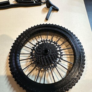 May include: A black bicycle wheel with a detailed spoke and gear design. The tire has a textured surface. Additional black bicycle parts are scattered around the wheel, including a seat and other components.
