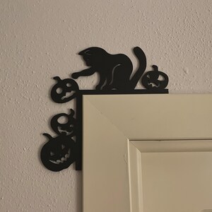 May include: A black metal door corner decoration featuring a silhouette of a cat reaching towards a small pumpkin, with two additional carved pumpkins positioned vertically along the side of the door frame, creating a festive seasonal accent.