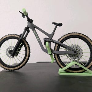 May include: A miniature gray mountain bike with black tires and light green accents. The bike has a black seat, handlebars, and suspension forks. The tires have a tan sidewall and the bike is supported by a light green stand.
