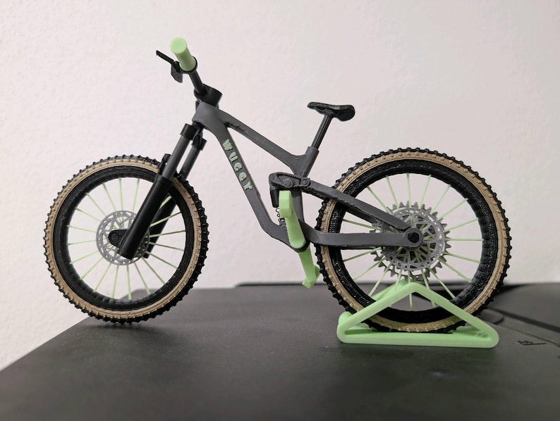 May include: A miniature gray mountain bike with black tires and light green accents. The bike has a black seat, handlebars, and suspension forks. The tires have a tan sidewall and the bike is supported by a light green stand.
