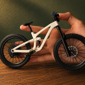 May include: A miniature white and black mountain bike with detailed components, including knobby tires, suspension, and a small seat. The bike is held in a hand, showcasing its size and design.