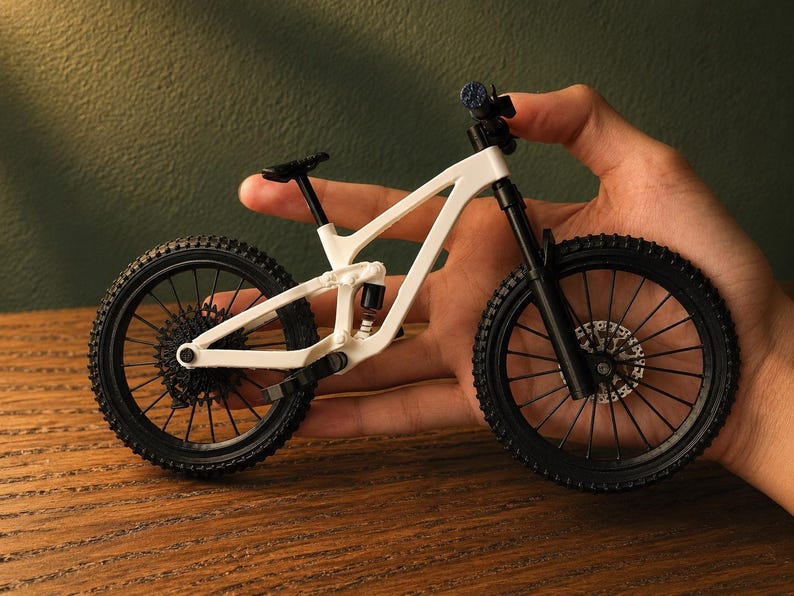 May include: A miniature white and black mountain bike with detailed components, including knobby tires, suspension, and a small seat. The bike is held in a hand, showcasing its size and design.