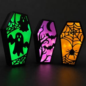 May include: Three coffin-shaped light boxes with black frames. The left box glows green with ghost and spiderweb silhouettes. The middle box glows purple with bat and tree silhouettes. The right box glows orange with a spider and web silhouette.