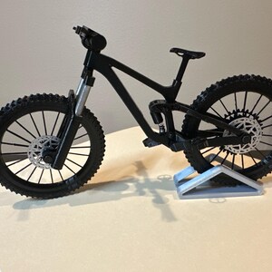 May include: A black mountain bike model with knobby tires and silver disc brakes. The bike is displayed on a light gray stand, showcasing its suspension and frame design. The model is a detailed representation of a full-suspension mountain bike.