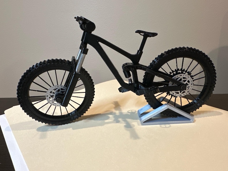May include: A black mountain bike model with knobby tires and silver disc brakes. The bike is displayed on a light gray stand, showcasing its suspension and frame design. The model is a detailed representation of a full-suspension mountain bike.