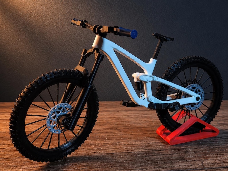 May include: A white and black mountain bike model with a blue handlebar grip. The bike has black tires, a black seat, and a red stand. The bike's frame is white, and the wheels have silver disc brakes.