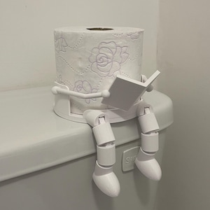 May include: A novelty white toilet roll holder. The holder features a roll of floral-patterned toilet paper and a white figure reading a book while seated on the toilet. The figure's legs are dangling.
