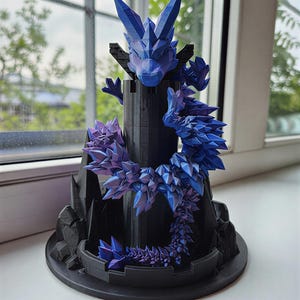 May include: A 3D-printed dragon sculpture in shades of blue and purple, perched atop a black castle-like structure. The dragon's body and the castle are intricately detailed, creating a fantasy-themed decorative piece.