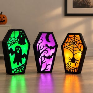 Puede incluir: Tres cajas de luz con forma de ataúd y marcos negros. La caja de la izquierda brilla en verde con siluetas de fantasmas y telarañas. La central brilla en morado con murciélagos y árboles. La derecha brilla en naranja con una araña.
