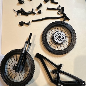 May include: Black mountain bike parts are arranged on a light brown surface. The components include two wheels with black tires and silver disc brakes, a frame, and various other parts. The bike appears to be disassembled.