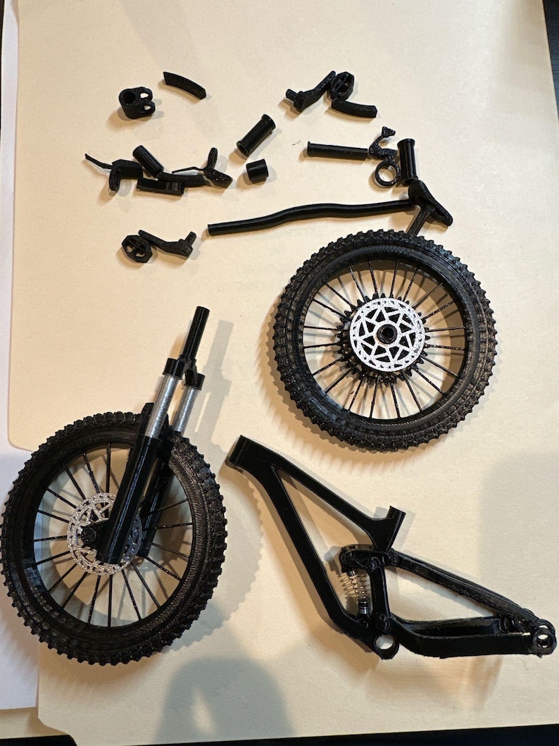 May include: Black mountain bike parts are arranged on a light brown surface. The components include two wheels with black tires and silver disc brakes, a frame, and various other parts. The bike appears to be disassembled.