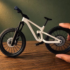 May include: A miniature white mountain bike with black tires and components. The bike features a suspension system, disc brakes, and a black seat. The handlebars have blue grips. The bike is displayed on a wooden surface.