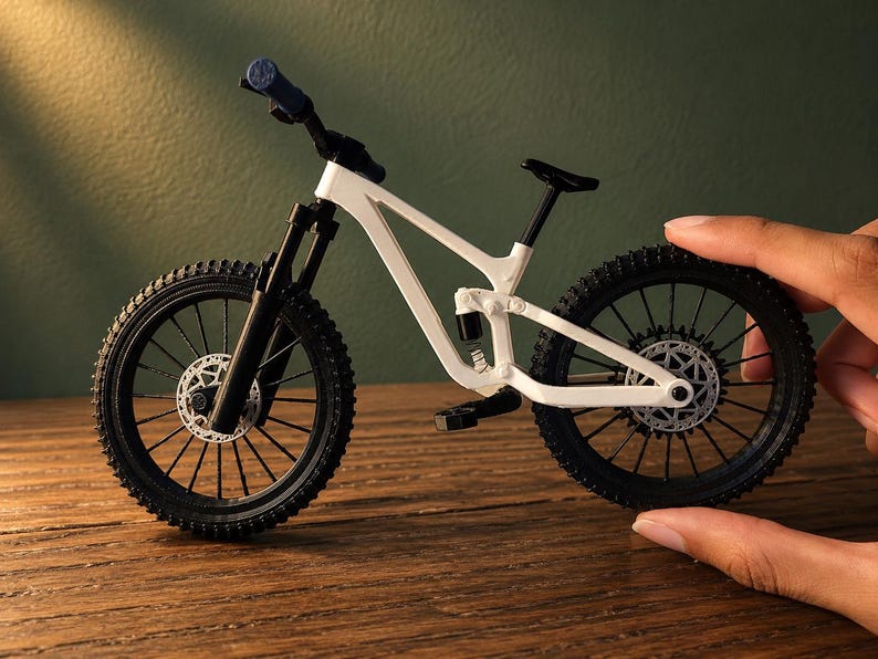 May include: A miniature white mountain bike with black tires and components. The bike features a suspension system, disc brakes, and a black seat. The handlebars have blue grips. The bike is displayed on a wooden surface.