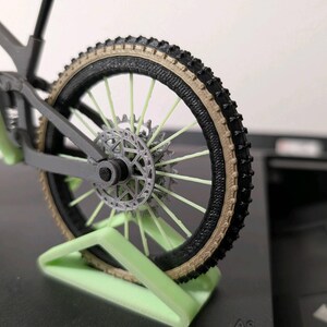 May include: A miniature gray mountain bike model with black tires and a tan sidewall. The bike features a disc brake and is displayed on a light green stand. The spokes are a light green color.
