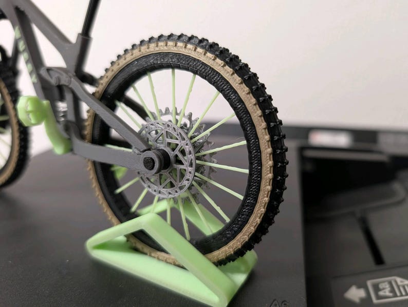 May include: A miniature gray mountain bike model with black tires and a tan sidewall. The bike features a disc brake and is displayed on a light green stand. The spokes are a light green color.