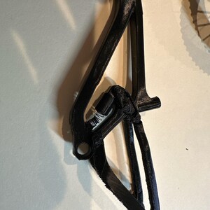 May include: A black bicycle frame component with a complex, angular design. The component has multiple connecting points and a textured surface, suggesting a 3D-printed or cast construction. The overall shape is elongated and features several holes for mounting.