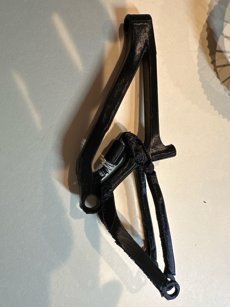 May include: A black bicycle frame component with a complex, angular design. The component has multiple connecting points and a textured surface, suggesting a 3D-printed or cast construction. The overall shape is elongated and features several holes for mounting.