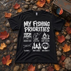 Puede incluir: Una camiseta negra de manga corta con el texto en blanco MY FISHING PRIORITIES y seis iconos gráficos con las etiquetas ORGANIZE GEAR, CATCH FISH, CAMP LAKESIDE, TIE KNOTS, SWAP STORIES y BRAG ABOUT IT, que representan diversas actividades de pesca.