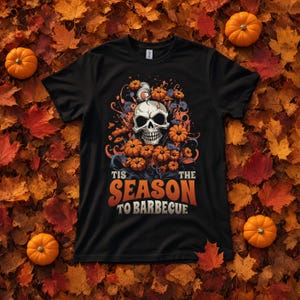 Puede incluir: Una camiseta negra de manga corta con un gráfico de una calavera rodeada de calabazas naranjas y remolinos azules. Debajo de la ilustración, el texto 'TIS THE SEASON TO BARBECUE' aparece en una fuente retro llamativa de color naranja y blanco.