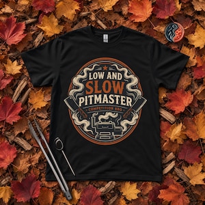 May include: A black short-sleeve t-shirt featuring a circular graphic with the text LOW AND SLOW PITMASTER COMPETITION BBQ. The design includes illustrations of two meat cleavers, a smoker grill, and swirling smoke patterns in orange and cream.