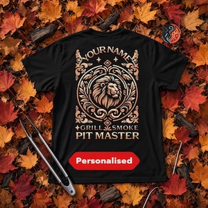 May include: A black t-shirt featuring a gold-toned graphic of a lion head inside an ornate circular frame. The text reads YOUR NAME, GRILL SMOKE PIT MASTER, and a red rectangular label with the word Personalised is printed below the design.