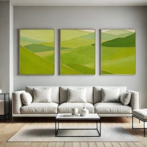 May include: Three framed abstract landscape paintings in shades of green with gold accents. The artwork depicts rolling hills and fields. The paintings are displayed above a white sofa in a modern living room.