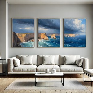 Puede incluir: Tres pinturas de paisajes enmarcadas representan acantilados costeros y olas del océano bajo cielos dramáticos y nublados. La obra de arte presenta una paleta de colores de azules, marrones y blancos, creando una atmósfera serena pero poderosa. Las pinturas se exhiben sobre un sofá blanco.