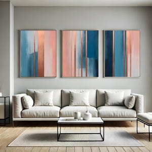 May include: Three framed abstract paintings with a modern design. Each artwork features vertical stripes in shades of blue, coral, and copper. The paintings are displayed above a white sofa in a living room setting.