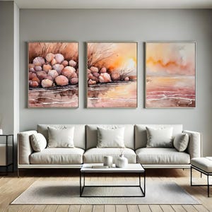 May include: Three framed watercolor paintings depicting coastal scenes. The artwork features a palette of warm colors, including shades of peach, brown, and orange. The subjects include seashells, water reflections, and a sunset over the ocean. The paintings are displayed above a white sofa.