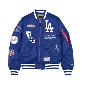 May include: A royal blue bomber jacket featuring white Los Angeles Dodgers embroidery, including the LA logo, team name, and various baseball-themed patches. The jacket has a front zip, striped ribbed cuffs and hem, and a red Alpha Industries tag.