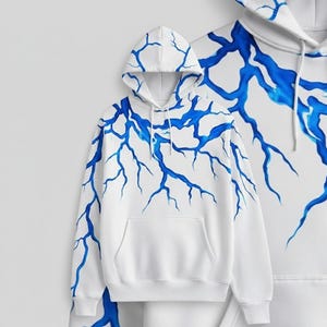 Op de afbeelding: Witte hoodie met een blauw bliksemontwerp. De hoodie heeft een capuchon met trekkoord en een voorzak. Het blauwe bliksemontwerp is geschilderd op de voorkant, capuchon en mouwen.