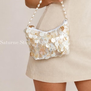 May include: A small handbag covered in iridescent, circular white and cream shell-like discs with a matching beaded handle. The bag features a zip closure and the text Saturne Studio is printed in white on the lower left side of the bag.