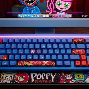 Può includere: Una tastiera meccanica blu con tasti rossi e personalizzati è posizionata su un tappetino per mouse di Poppy Playtime. La tastiera ha una barra luminosa ed è accanto a un monitor di computer che mostra i personaggi di Poppy Playtime. Sono visibili anche un mouse nero e delle cuffie blu.