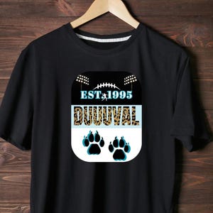 Puede incluir: Camiseta negra con un diseño gráfico que presenta un balón de fútbol, luces de estadio y el texto "EST 1995 DUUUVAL". Debajo del texto hay dos huellas de patas en verde azulado y negro. La camiseta está colgada en una percha de madera.