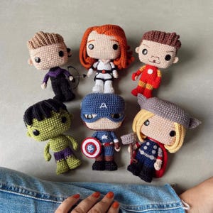 May include: Six handmade crochet dolls representing superhero characters. The collection includes figures with distinct features like a green body, a red and gold suit, a blue suit with a shield, a winged helmet, red hair, and a black outfit with a bow.